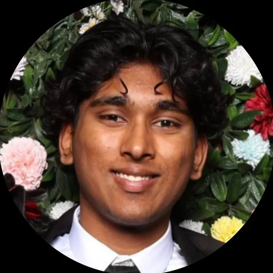 Sasank Parimi, smiling, in a dark jacket and tie in front of a wall of flowers and leaves.
